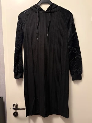 Svart hoodieklänning med spetsärmar - Svart långärmad hoodieklänning med huva och snörning. Ärmarna är dekorerade med spetsdetaljer som ger en snygg kontrast till den ribbade strukturen på resten av klänningen. Bara tagen ur förpackning för att fota, så helt ny.