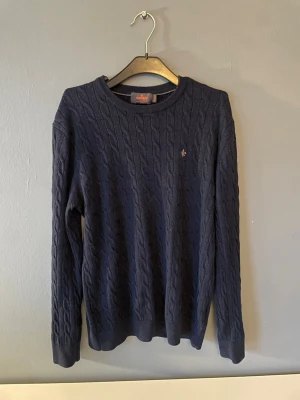 Mörkblå kabelstickad tröja Morris - Mörkblå kabelstickad tröja från Lexington med rund halsringning och diskret logga broderad på bröstet. Tröjan har långa ärmar och ribbade muddar vid ärmslut och nederkant. Perfekt för lager på lager och klassisk stil. Nypris 1200. Mitt pris 549!