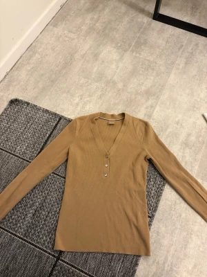 Beige ribbad topp från Boss - Snygg beige ribbad topp från Boss med lång ärm och djup V-ringning. Toppen har tre dekorativa knappar framtill och är tillverkad i ett stretchigt, mjukt material som sitter skönt på kroppen. Perfekt att styla med jeans eller kjol.