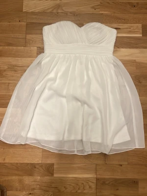 Vit axelbandslös kort klänning - Söt vit axelbandslös kort klänning med omlott över bysten och markerad midja. Kjolen är lätt utställd med ett tunt, svepande yttertyg som ger en luftig känsla.