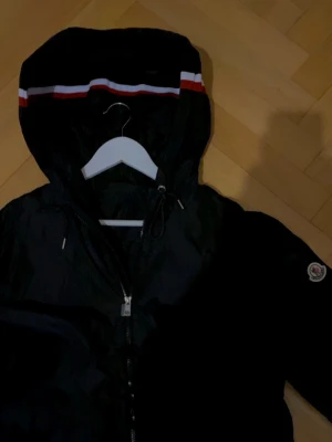 Svart Moncler vindjacka med huva ( NY ) - Moncler Grimpeus. Nyskick helt oanvänd. Snygg svart vindjacka från Moncler med huva som har röd och vit rand. Jackan har dragkedja framtill och Moncler-logga på ärmen. Perfekt för dig som vill ha en stilren och sportig look.