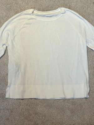 Vit stickad tröja med rund hals - En enkel och stilren vit stickad tröja med rund halsringning och långa ärmar. Tröjan har ribbstickad kant nedtill och är tillverkad i ett mjukt, tunt material. Längden är cirka 47 cm.