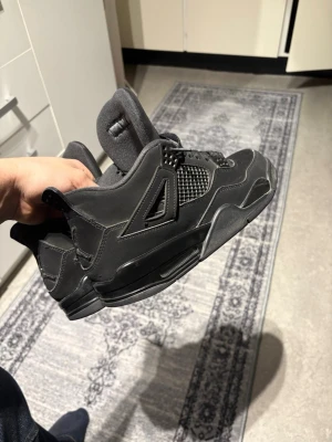Jordans 4 black cats - Säljer ett par helsvarta Nike Jordan 4 sneakers med meshdetaljer på sidorna, klassisk snörning och den ikoniska Jumpman-loggan på sulan. Skorna har en robust och sportig design med rund tå och platt sula. Perfekta för dig som gillar streetwear och snygga detaljer. Har haft dessa skor snart 1 månad. Inga defekter eller repor. Bara seriösa kunder.