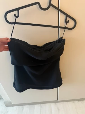 Svart axelbandslös topp med drapering - Snygg svart axelbandslös topp med draperad framsida och smala axelband. Toppen har en elegant look och dragkedja i sidan för enkel påklädning. Perfekt till ett par jeans eller kjol för en trendig stil.