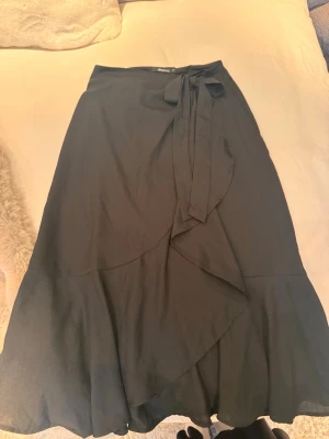 Svart omlottkjol med knytband från Bikbok  - Säljer en svart långkjol i omlottmodell med knytband i midjan. Kjolen har en volangkant nedtill som ger ett snyggt fall och är perfekt för dig som gillar en enkel men stilren look. Passar till många olika outfits och är superbekväm.