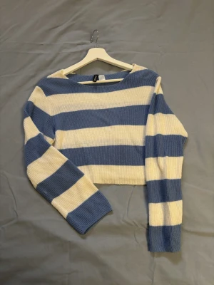 Blåvit randig cropped stickad tröja - Säljer en stickad cropped tröja med breda blå och vita ränder. Tröjan har långa ärmar och en avslappnad passform, perfekt för en trendig vardagslook. Materialet är mjukt och ribbstickat för extra komfort.