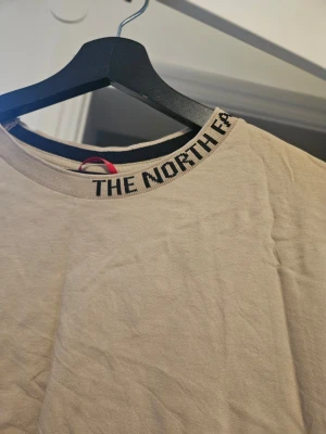 Beige croppad topp från The North Face - Säljer en beige croppad topp från The North Face med svart logotyptext runt halsringningen. T-shirten har korta ärmar och är tillverkad i mjuk bomull, perfekt för en avslappnad och trendig look.