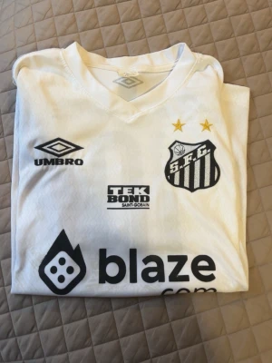 Vit Santos matchtröja Umbro - Santos FC matchtröja i vitt från Umbro med svarta tryck och flera sponsorer. Nummer 10 och namnet Santos på ryggen. Två gula stjärnor ovanför klubbmärket. Klassisk V-ringning och sportig passform, perfekt för fotboll eller samling.