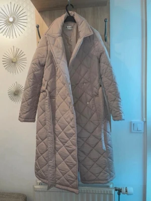 Beige quiltad lång pufferjacka - Lång beige pufferjacka med quiltat mönster och klassisk krage. Jackan har en rak passform, tryckknappar framtill och sidfickor. Perfekt för kyliga dagar och ger en clean, trendig look.