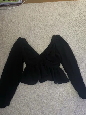 Svart topp från H&M med volang - Snygg svart omlottopp från H&M i storlek XS. Toppen har v-ringning, långa ärmar och volangdetalj i midjan som ger en feminin siluett. Perfekt att styla med både jeans och kjol för en trendig look.