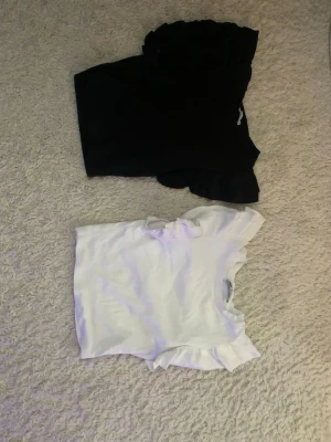 Svart och vit topp med volangärm - Två toppar i svart och vitt med breda volangärmar. Båda har rund halsringning och är tillverkade i mjukt material, perfekta för en trendig och stilren look. Passformen är normal och toppen är ärmlös med volangdetaljer som ger extra attityd.