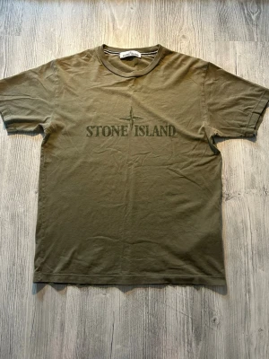Olivgrön Stone Island t-shirt - Snygg olivgrön t-shirt från Stone Island med klassisk logga tryckt på bröstet. T-shirten har rund hals och korta ärmar, perfekt för en avslappnad och trendig stil. Tillverkad i mjuk bomull som känns skön mot huden. Äkta