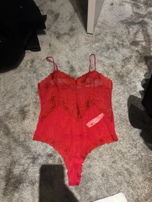 Röd spetsbody med tunna axelband - Snygg röd body i spets med tunna axelband och dekorativa spetsdetaljer. Bodyn har v-ringad urringning, genomskinligt tyg och tryckknappar i grenen. Perfekt för dig som vill ha något extra under kläderna. Helt oanvänd! Nypris 600!
