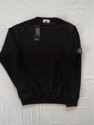 Svart sweatshirt från Stone Island - Säljer en svart sweatshirt från Stone Island med klassisk rund hals och ribbade muddar. Tröjan har den ikoniska Stone Island-loggan på vänster ärm och är tillverkad i mjukt bomullsmaterial. Perfekt för en clean och stilren look.