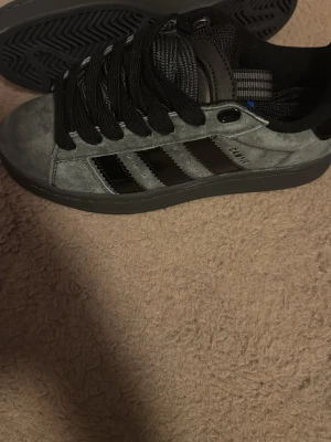 Adidas Campus grå/svarta sneakers - Snygga Adidas Campus sneakers i grå mocka med svarta läderdetaljer och klassiska tre ränder på sidan. Skorna har svart platt sula, rund tå och snörning. Perfekta för dig som gillar stilrena och tidlösa sneakers.