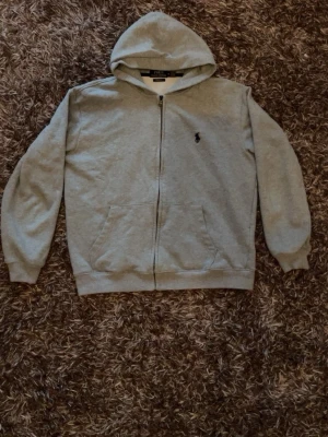 Grå hoodie från Polo Ralph Lauren - Klassisk grå hoodie med dragkedja från Polo Ralph Lauren. Hoodien har två fickor framtill, broderad logga på bröstet och mjukt bomullsmaterial. Perfekt för en avslappnad och stilren look.