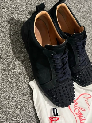 Mörkblå Christian Louboutin sneakers med spikar. - Säljer ett par mörkblåa sneakers från Christian Louboutin i mocka med coola med spikar. Skorna har snörning, rund tå och klassisk röd sula. Insidan är i ljust skinn och de kommer med original dustbag. Perfekta för dig som vill sticka ut.