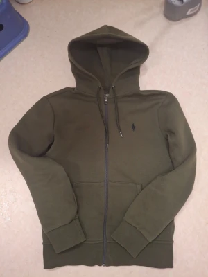 Olivgrön zip hoodie från Polo Ralph Lauren - Snygg olivgrön zip hoodie från Polo Ralph Lauren med dragkedja framtill, två fickor och klassisk huva med dragsko. Liten broderad logga på bröstet. Tillverkad i mjuk bomullsblandning som känns skön mot huden. Perfekt för en avslappnad och stilren look. Kvittot följer med om du vill!
