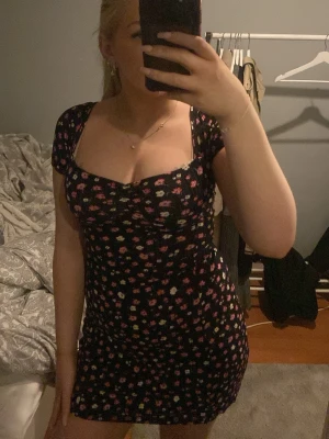 Svart blommig kortklänning med snörning - Snygg svart kortklänning med färgglatt blommönster och snörning i ryggen. Säljer pågrund av att den är för liten och för att jag inte får någon användning för den