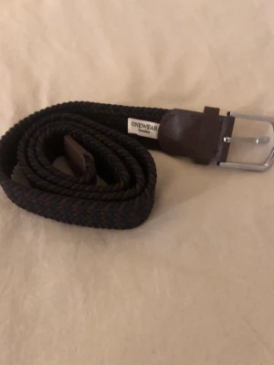 Onewear Skärp  - Bilderna visar ett flätat bälte från ONEWear Sweden i mörkbrunt och svart med detaljer i läder och metallspänne. Storlek 100. Köptes till lillebrorsan men tyvärr var det fel storlek