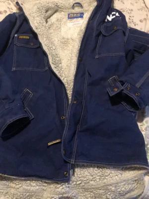 Blå teddyjacka från Blåkläder - Mysig blå teddyjacka från Blåkläder med vit fluffig insida. Jackan har flera fickor, synliga sömmar och stängs med knappar. Perfekt för kalla dagar när du vill vara både varm och snygg.