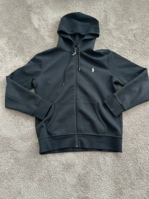 Svart hoodie från Polo Ralph Lauren - Klassisk svart hoodie med dragkedja från Polo Ralph Lauren. Tröjan har huva med dragsko, två fickor framtill och den ikoniska lilla loggan broderad på bröstet. Vid frågor är det bara att slänga iväg ett meddelande!