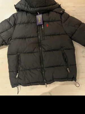 Svart pufferjacka från Polo Ralph Lauren - Snygg svart pufferjacka från Polo Ralph Lauren med röd broderad logga på bröstet. Jackan har dragkedja framtill, två sidofickor med dragkedja och huva. Perfekt för kalla dagar och riktigt trendig look. Storlek M, nytt skick.