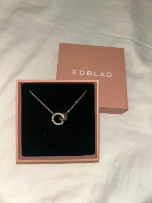 Silverhalsband med cirkel från Edblad - Snyggt halsband från Edblad i silverfärg med två sammanlänkade cirklar, varav en är dekorerad med små gnistrande stenar. Kedjan är tunn och elegant, perfekt för att lyfta vilken outfit som helst. Kommer i original ask och har aldrig använt.