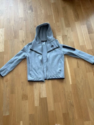 Nike tech fleece - Snygg grå hoodie från Nike i storlek 137-147  med hel dragkedja framtill och två sidofickor. Hoodien har svarta detaljer, inklusive dragkedjor och en cool ficka på ena ärmen. Tillverkad i mjukt material och har en klassisk Nike-logga på bröstet. Perfekt för en sportig och avslappnad stil. 