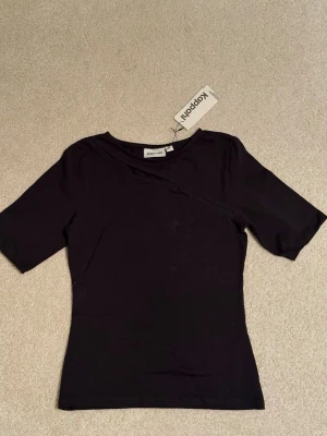 Svart topp från Kappahl - Snygg svart topp från Kappahl med korta ärmar och rund halsringning. Toppen har en diskret asymmetrisk snitt framtill som ger en cool detalj. Ny med prislapp.
