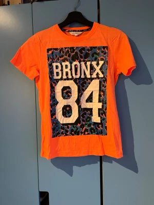 Orange t-shirt  - Cool orange t-shirt från KappAhl med stort tryck 'BRONX 84' på bröstet och blått leopardmönster i bakgrunden. Klassisk rund hals och korta ärmar. Perfekt för dig som gillar streetstyle och färgstarka plagg.
