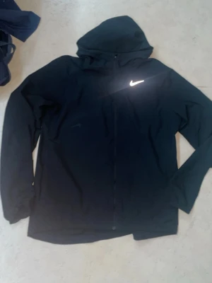 Svart Nike Dri-FIT vindjacka M - Säljer en svart Nike Dri-FIT vindjacka med huva och hel dragkedja. Jackan har en clean look med reflekterande Nike-logga på bröstet och är tillverkad i lätt polyester som andas. Perfekt för löpning eller utomhusaktiviteter. Passar S oxå