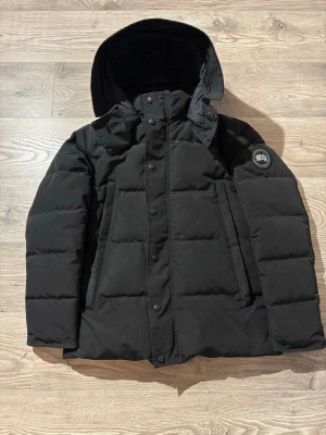 Canada Goose - Säljer en svart dunjacka från Canada Goose med black label, huva och klassisk patch på ärmen. Jackan har quiltad design, dragkedja och knappar framtill samt två stora fickor. Perfekt för kalla vinterdagar och riktigt snygg streetstil.