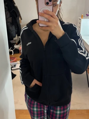 Svart Adidas hoodie med dragkedja - Svart hoodie från Adidas med klassiska vita ränder längs ärmarna och vit Adidas-logga på bröstet. Dragkedja framtill, huva och två fickor. Perfekt för en sportig och avslappnad stil.