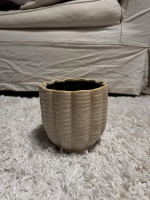 Beige räfflad kruka i keramik - Säljer en beige kruka i keramik med  vågig kant upptill. Krukan har en rundad form och passar perfekt för växter eller som dekoration. Den neutrala färgen gör att den smälter in i de flesta inredningsstilar.