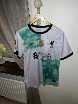 Fotbollströja - Säljer en Liverpool FC bortatröja från Nike med Virgil 4 på ryggen. Tröjan är vit med gröna grafiska mönster, svarta detaljer vid krage och ärmslut samt officiella klubb- och sponsorloggor på bröstet. 
