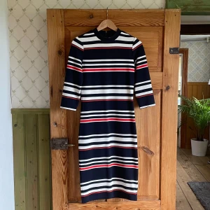 Randig marinblå klänning från Yessica - Snygg randig klänning från Yessica i marinblått, vitt och rött. Modellen är knälång med trekvartsärm och har en smickrande passform. Klänningen har dragkedja i ryggen och är gjord i mjukt, stretchigt material som känns bekvämt hela dagen.