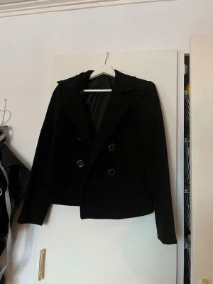 Svart kort kappa - Stilren svart kappa med dubbelknäppning och klassisk krage. Perfekt för en trendig och clean look.