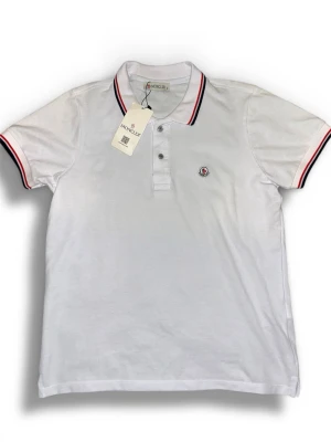 Vit Moncler pikétröja  - Klassisk vit pikétröja från Moncler med korta ärmar och krage. Tröjan har tunna röda och marinblå ränder på krage och ärmslut samt Moncler-logga på bröstet. Perfekt till jeans eller shorts för en clean och sportig look.