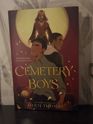 Cemetery Boys av Aiden Thomas (Hardcover) - En skönlitterär bok med hårt omslag i vinrött och guld, med illustration av tre personer mot en gul måne. Omslaget har guldtext och blommotiv på insidan. Boken är på engelska och blandar magi, identitet och spänning.