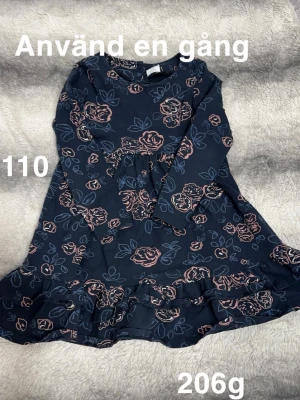 Mörkblå blommig klänning med volang - Mörkblå klänning med rosa blommönster och volangkant nedtill. Långärmad modell i mjukt material, perfekt för en trendig vardagslook. 🌸 Använd en gång. Storlek 110 