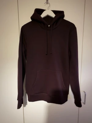 Vinröd hoodie - En stilren mörkbrun hoodie med huva och dragsnören. Hoodien har en klassisk känguruficka framtill och ribbade muddar vid ärmar och nederkant. Perfekt för en avslappnad och trendig look. Materialet känns mjukt och bekvämt.