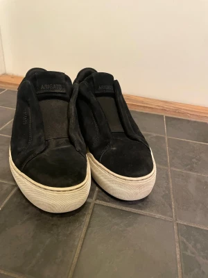 Svarta sneakers från Axel Arigato laceless  - Stilrena svarta sneakers från Axel Arigato med ovandel i mocka och bred resår över vristen. Skorna har en vit platt sula och minimalistisk design utan snörning. Perfekta för en clean och modern look.