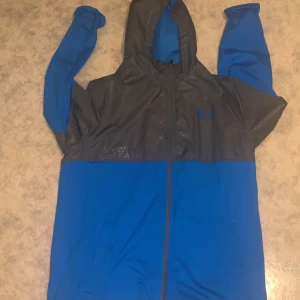 Blå och grå vindjacka från Under Armour - Snygg vindjacka från Under Armour i blått och mörkgrått. Jackan har huva, hel dragkedja framtill och långa ärmar. Perfekt för blåsiga dagar och sportiga outfits. Tillverkad i lätt polyester som andas och ger bra rörelsefrihet.