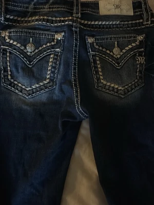 Miss Me bootcut jeans med broderi - Snygga mörkblå jeans med bootcut passform och kontrastsömmar. Bakfickorna har detaljerat broderi och dekorativa knappar. Jeansen har låg midja och är tillverkade i klassiskt denimtyg. Längden passar för de som är runt 162-168. De är använda 2-3 ggr då jag tycker att de är för korta för mig som är 173