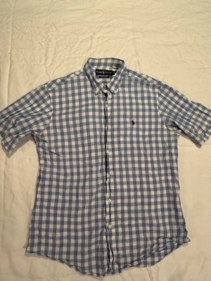 Blårutig skjorta från Ralph Lauren - Klassisk blårutig skjorta från Ralph Lauren med korta ärmar och button-down krage. Skjortan har broderad logga på bröstet och är tillverkad i bomull. Custom fit-modell som ger en snygg passform. Perfekt för en avslappnad men stilren look.