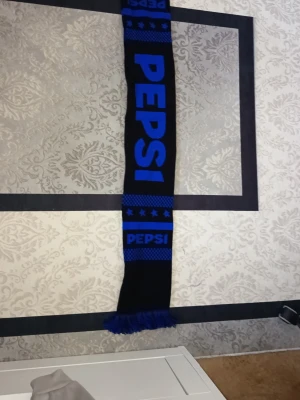 Blå och svart Pepsi halsduk - Säljer en cool halsduk med Pepsi-logga i blått på svart bakgrund. Halsduken har fransar i ändarna och stjärnmönster upptill och nedtill. Materialet är 100% akryl och den är tillverkad i EU. Perfekt för dig som vill sticka ut med en unik accessoar. Limited Edition pepsi halsduk.