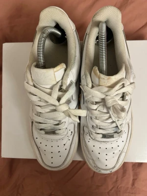 Nike Air Force 1 vita sneakers - Klassiska Nike Air Force 1 sneakers i helvitt skinn med perforerad tå, rund tå och vit sula. Skorna har snörning och den ikoniska swoosh-loggan på sidan. Låg modell som passar till det mesta och har en tidlös streetstil. (Ingen box)