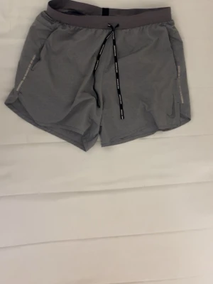 Grå träningsshorts från Nike - Säljer ett par grå Nike träningsshorts med svart snörning där det står 'NIKE RUNNING'. Shortsen har en loose passform, midwaist och är tillverkade i ett lätt syntetmaterial. Swoosh-loggan syns på ena benet och de har praktiska fickor på sidorna.