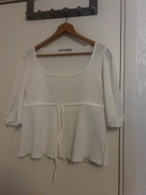 Vit blus med knyt från 157 - Supersöt vit blus från 157 med fyrkantig ringning och trekvartsärm. Blusen har ett tunt ribbat tyg i bomull och ett knytband i midjan som ger en fin siluett. Perfekt till jeans eller kjol för en fräsch look.